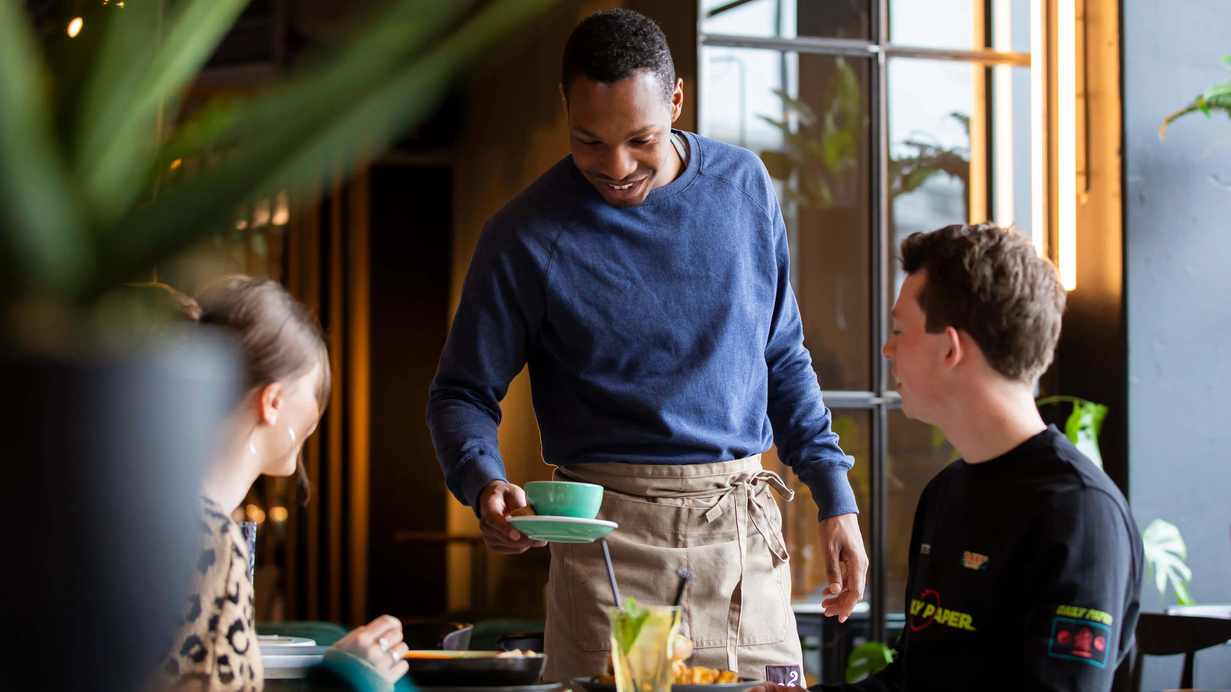 Medewerker bediening serveert een kopje koffie aan twee gasten. bediening, horeca, hai