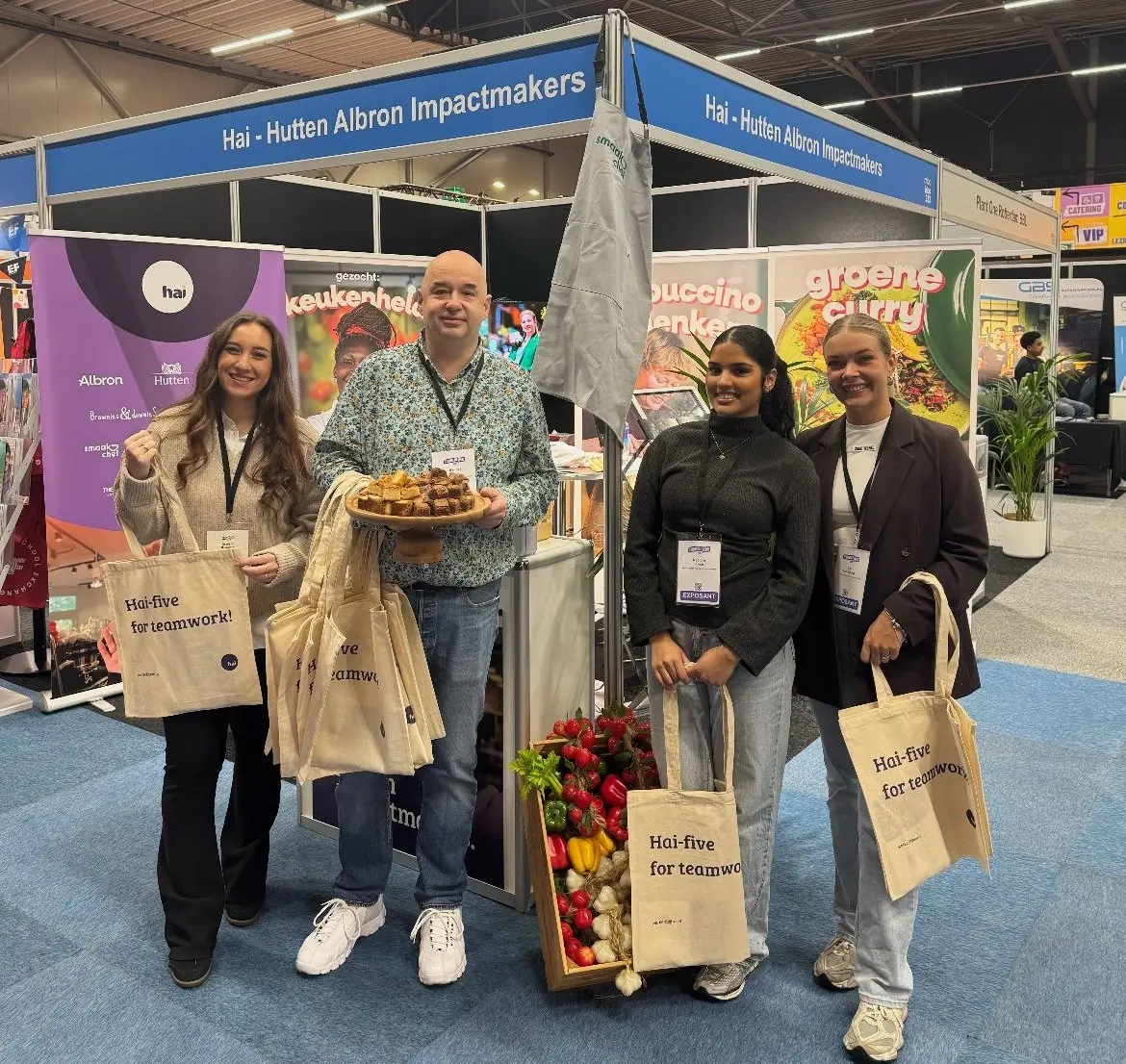 Vier personen staan bij een beursstand met een blauwe banner waarop staat: “Hai – Hutten Albron Impactmakers”. De stand is kleurrijk ingericht met posters van gerechten zoals. Voor de stand staat een houten krat gevuld met verse groenten. Eén persoon houdt een schaal met brownies, terwijl de anderen stoffen tassen vasthouden met de tekst: “Hai-five for teamwork!”.
studiekeuzebeurs, Hai
