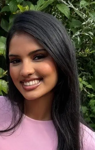 Een persoon draagt een lichtroze shirt met een ronde hals. Het haar is lang, steil en donker, en valt over één schouder naar voren. De achtergrond bestaat uit groene bladeren en planten, wat een natuurlijke, buitenomgeving suggereert. Hai, Hutten, Albron,