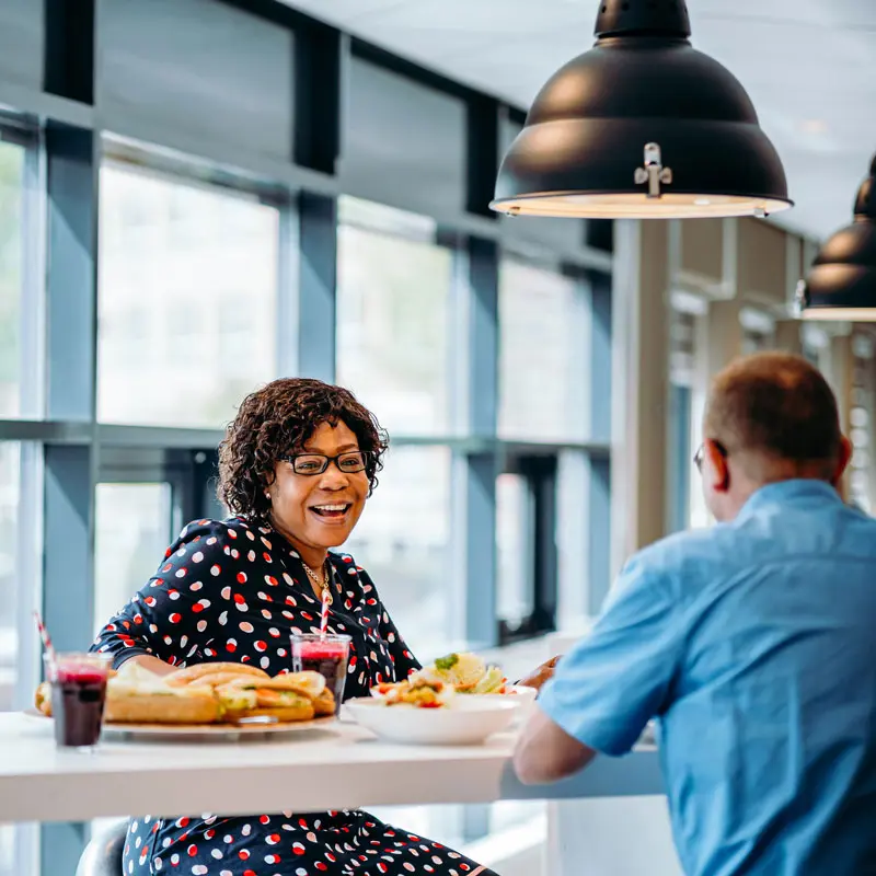 Twee personen zitten aan een hoge witte tafel in een moderne, lichte ruimte met grote ramen. Op de tafel staan borden met broodjes, een salade en glazen met rode drank en rietjes. Boven de tafel hangen grote zwarte industriële lampen. Servicekantoor, Hai