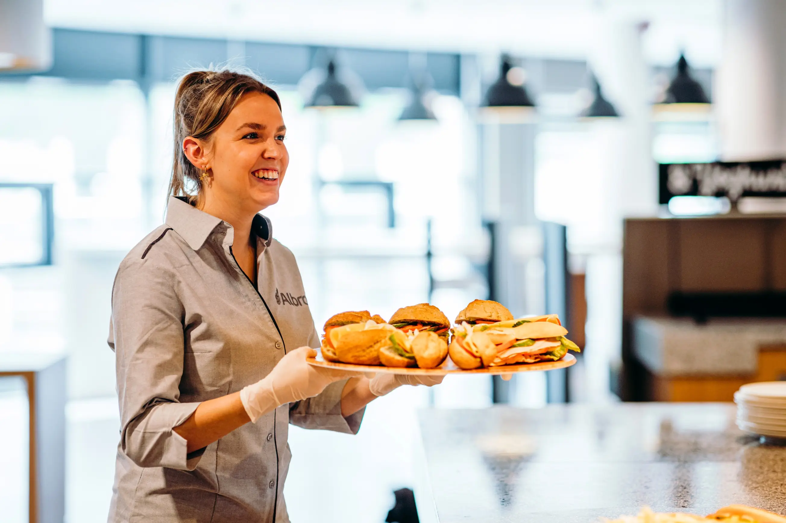 Een medewerker draagt een lichtgrijze blouse met het logo “Albron” op de borst en witte handschoenen. De medewerker houdt een groot, rond dienblad vast met meerdere belegde broodjes. De broodjes zijn divers gevuld, onder andere met sla, kaas en vleeswaren. Hai, Lunch